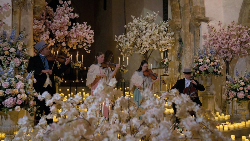 Musicians playing string instruments in a decorated indoor setting with floral arrangements and candles.