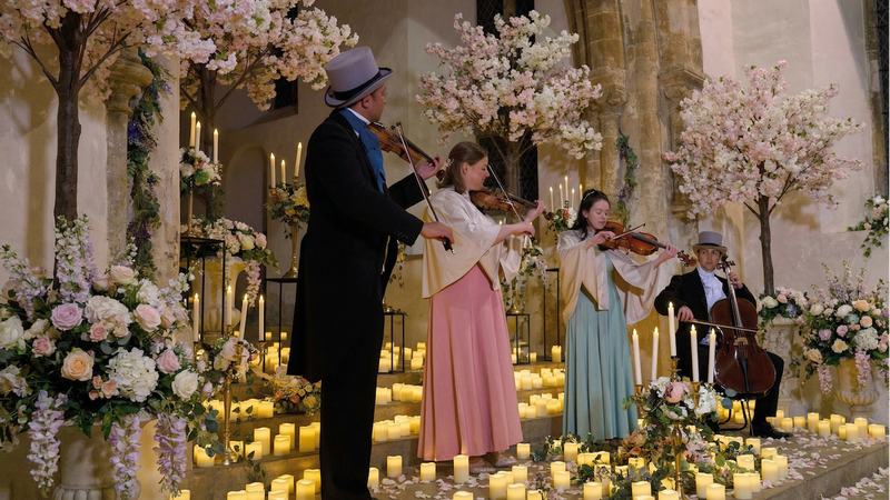 Four musicians playing string instruments in a decorated indoor setting with flowers and candles.