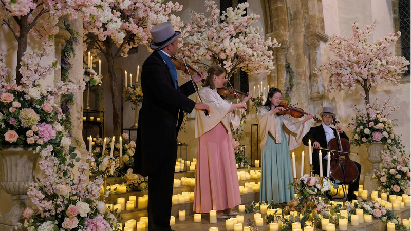 Four string musicians performing as a quartet in a floral, Regency-style environment with an elegant, classical atmosphere.
