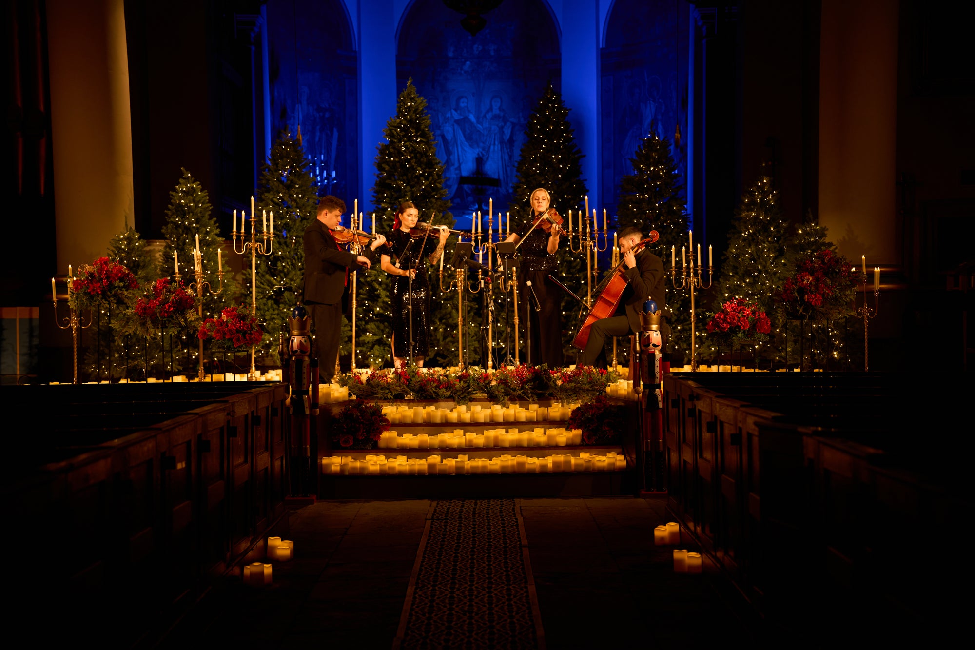 A festive holiday concert scene in a church or hall, with four musicians performing on a raised stage decorated with lit candles, small Christmas trees, and red floral arrangements. The background is dimly lit with blue ambient lighting.