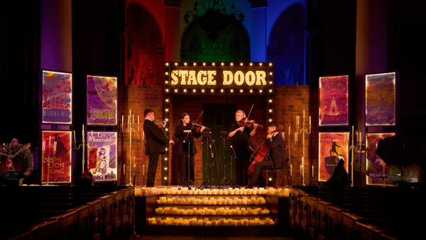 A live orchestral performance on a dimly lit stage with a bright 'STAGE DOOR' marquee sign, musicians playing string instruments, and colorful theatrical lighting in purple, green, and red.