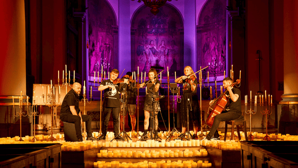 A group of five musicians performing on a stage inside a church or cathedral, surrounded by numerous lit candles. The background features ornate arched wall panels with painted religious scenes illuminated by purple and warm orange lighting.