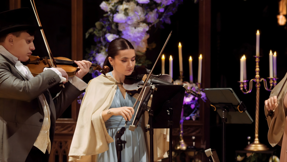 Two musicians playing violins in a dimly lit setting with candles and decorative elements.
